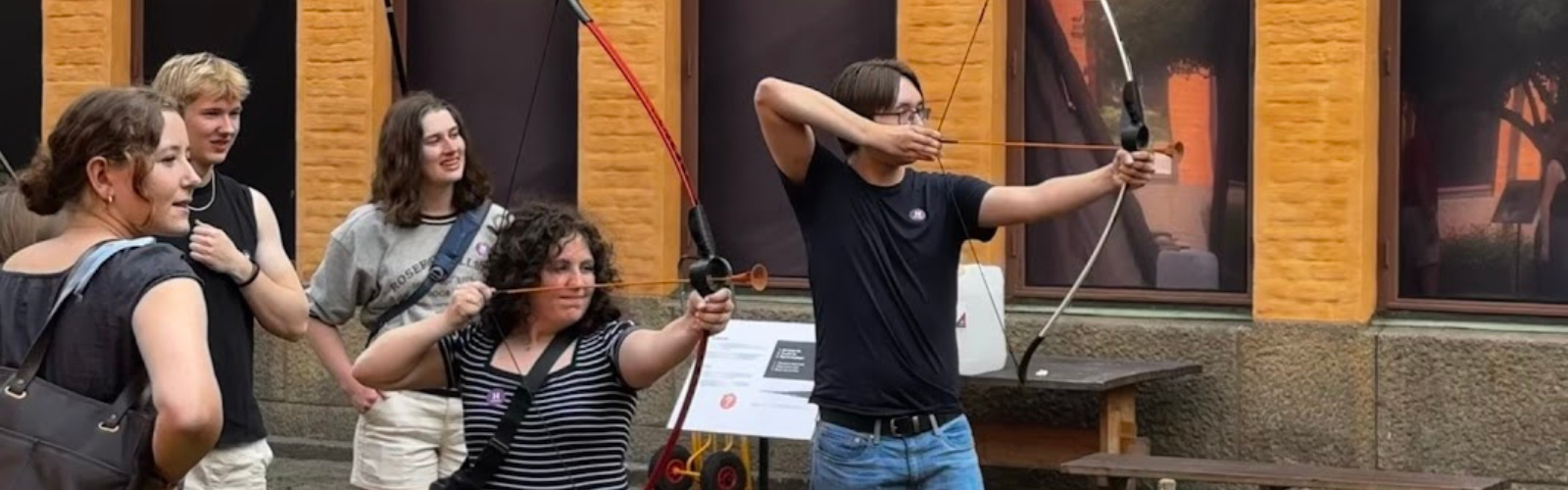 Two students aiming bows and arrows (with soft tips) while three other students watch from behind.